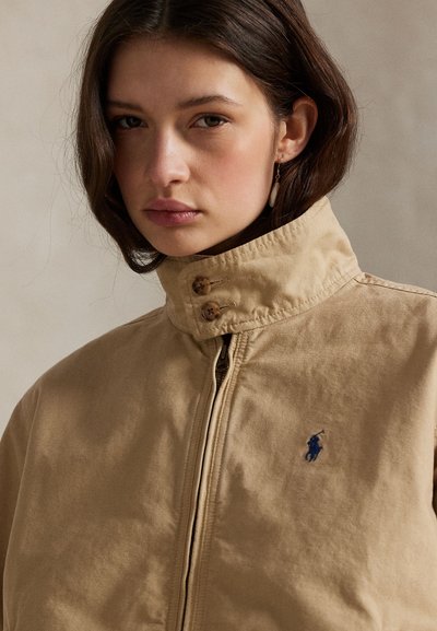 Ung kvinne med brunt hår iført beige glidelåsjakke med høy krage og liten brodert blå logo på brystet, nøytral bakgrunn.