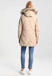 Parka beige con cappuccio foderato in pelliccia, texture trapuntata e lunghezza lunga, abbinata a jeans blu e sneakers bianche.