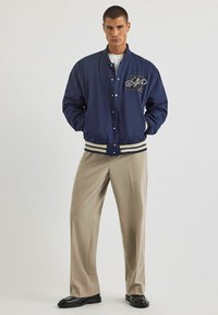 Granatowa kurtka bombowca z ściągaczami przy mankietach i dolnej krawędzi, z ozdobną naszywką na lewej piersi. Noszona na beżowych, dopasowanych spodniach.