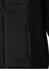 Gros plan d'une veste noire avec un panneau avant matelassé, fermeture éclair et poche latérale zippée, montrant la texture du tissu et les détails des coutures.