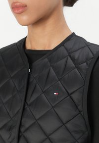 Černá prošívaná vesta s kulatým výstřihem a kontrastním lemováním. Na hrudi se nachází malé značkové logo v červené, bílé a modré barvě.