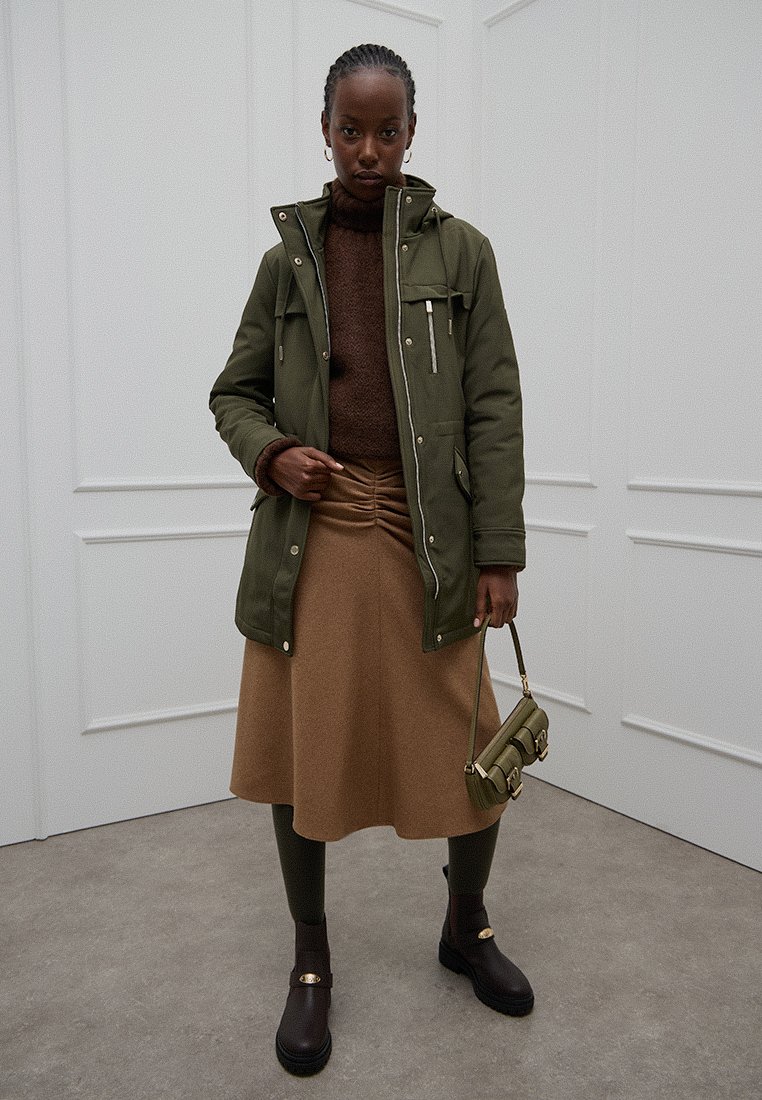 Parka verde oliva, dolcevita marrone a maglia, gonna marrone, collant verdi, stivali marroni chunky con dettagli dorati e una piccola borsa verde.