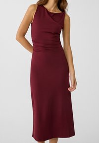 Bordeaux mouwloze jurk met een gedrapeerde halslijn, subtiele plooien bij de taille en een lengte tot halverwege het scheenbeen, gemaakt van een gladde, rekbare stof.