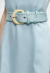 Φωτεινά γαλάζια υφασμάτινη ζώνη με μεγάλο μπεζ υφαντό αγκράφα, φορεμένη πάνω από ασορτί φωτεινά γαλάζιο φόρεμα.