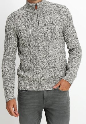 Sweter dziany w czarno-białym melanżowym wzorze, z wysokim kołnierzem i zamkiem, z ribbingu przy mankietach i u dołu. Noszony z szarymi spodniami.