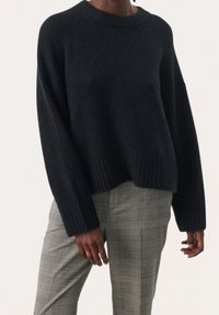 Personne portant un pull noir texturé avec des poignets côtelés et un pantalon à carreaux gris, debout devant un fond clair uni.
