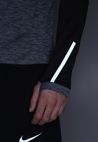 Schwarz und graues Sportshirt mit langen Ärmeln, das einen reflektierenden weißen Akzent entlang des Arms und ein strukturiertes Gewebe am Handgelenk aufweist.