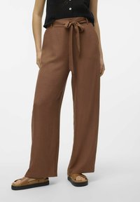 Pantalons marron à jambes larges en matériau texturé. Dotés d'une ceinture en tissu nouée à la taille. Portés avec des sandales marron laissant voir les orteils.