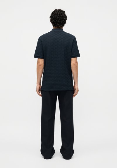 Άνδρας με σγουρά μαλλιά που φοράει ένα υφασμάτινο σκούρο κοντομάνικο πόλο και φαρδιά μαύρη παντελόνια, στέκεται με την πλάτη σε απλό λευκό φόντο.