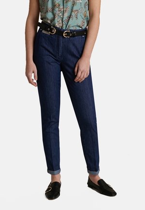 Femme portant un jean bleu foncé retroussé, une ceinture noire avec boucles dorées, un chemisier vert à motifs et des mocassins noirs avec détails de boucle dorée.