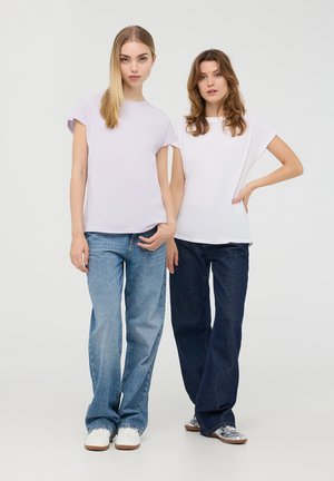 Due model indossano una camicia a maniche corte, una di colore lilla chiaro e l'altra bianca, abbinate a jeans a fit largo in blu chiaro e scuro.