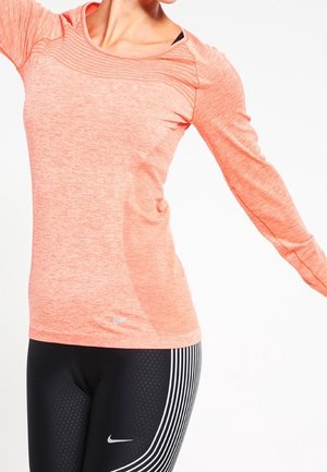 Kobieta w koralowej sportowej bluzce z długim rękawem i czarnych legginsach Nike z białymi paskami, z rozpostartymi ramionami na białym tle.