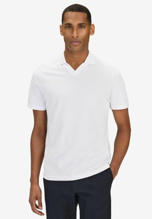 Mann in schlichter weißer kurzärmeliger Poloshirt und dunkler Hose, der mit einer Hand in der Tasche vor einfarbigem Hintergrund steht.