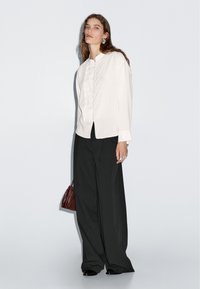 Witte jas met lange mouwen, een knoopsluiting aan de voorkant en een rucherandje. Gecombineerd met zwarte wijde broekspijpen. Accessoires omvatten een kleine bruine tas.