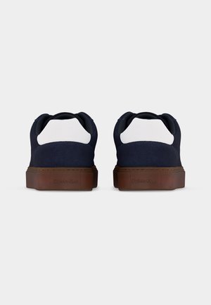 Paire de baskets basses bleu marine et blanches avec semelles marron vues de dos sur un fond gris clair uni.