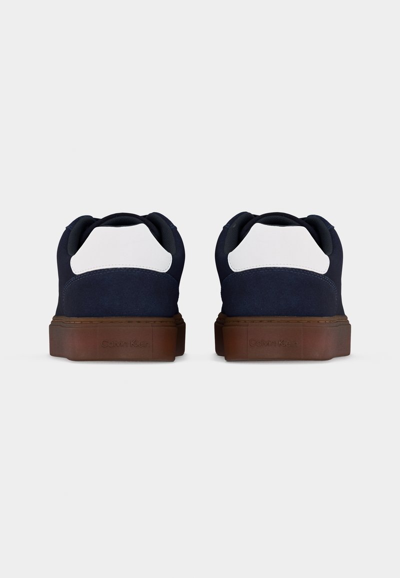 Paire de baskets basses bleu marine et blanches avec semelles marron vues de dos sur un fond gris clair uni.