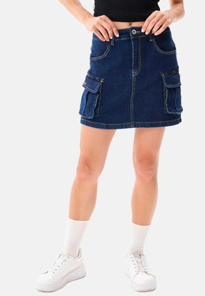Jupe en denim bleu foncé avec une coupe droite, fermeture par boutons à l'avant et deux grandes poches cargo latérales. Portée avec des baskets blanches et des chaussettes.