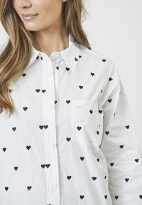 Blusa bianca con motivi a cuore ricamati neri, chiusura con bottoni, colletto a risvolto e taschino singolo sul petto, caratterizzata da una texture liscia.