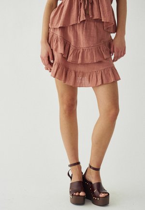 Kobieta ubrana w różową warstwową spódnicę z falbankami i brązowe sandały na platformie z paskami wokół kostek, stojąca na jednolitym tle.