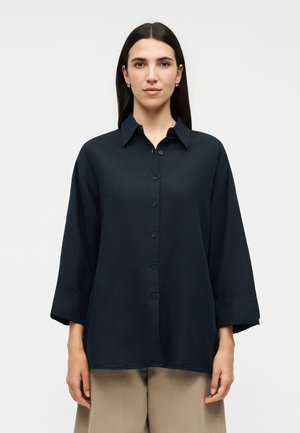 Γυναίκα με ίσια σκουρόχρωμα μαλλιά φοράει χαλαρό μαύρο πουκάμισο με κουμπιά και φαρδιά μανίκια και μπεζ παντελόνι, στέκεται μπροστά σε απλό ανοιχτό φόντο.