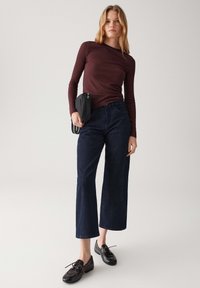 Donna in piedi con indosso un top a maniche lunghe bordeaux a costine, jeans scuri a gamba larga, scarpe nere con lacci e che tiene in mano una piccola borsa nera.