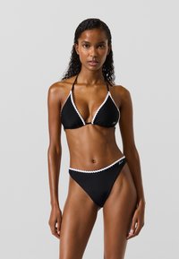 Set di bikini nero composto da un top triangolare con bordo ondulato bianco e slip a vita bassa coordinati. Tessuto liscio e testurizzato con spalline regolabili.