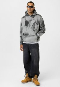 Grijze tie-dye hoodie met een voorzak, gedragen over een wit shirt, gecombineerd met donkere baggy jeans en gele werklaarzen. Zonnebril inbegrepen.