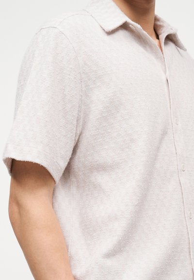 Άνδρας που φοράει ανοιχτό μπεζ κοντομάνικο πουκάμισο με κουμπιά, κατασκευασμένο από μαλακό, υφασμάτινο ύφασμα με υφή και διπλωμένο γιακά.