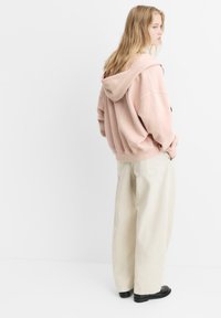 Hellrosa übergroße Kapuzenjacke mit Kängurutasche und Kapuze, kombiniert mit weiten beigen Hosen und schwarzen Schuhen, von hinten gezeigt.