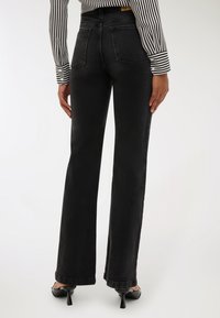 Kobieta w czarnych, wysokich jeansach o prostym kroju, czarnych szpilkach oraz czarno-białej, paskowanej koszuli z długim rękawem, stojąca.