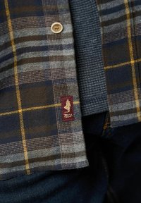 Karirana flanel košulja s plavim, sivim i žutim uzorcima; sadrži istaknutu vezenu oznaku u maroon boji i zlatni gumb na patentu.