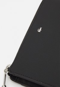 Portefeuille noir texturé avec un logo en crocodile argenté, fermeture éclair et accent en métal. Forme rectangulaire compacte avec des bords lisses.