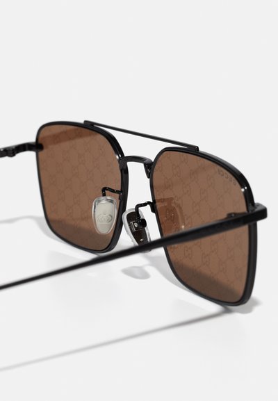 Gafas de sol cuadradas negras con lentes de color marrón, que presentan un sutil patrón de logo y acentos metálicos. Incluyen almohadillas ajustables para la nariz.