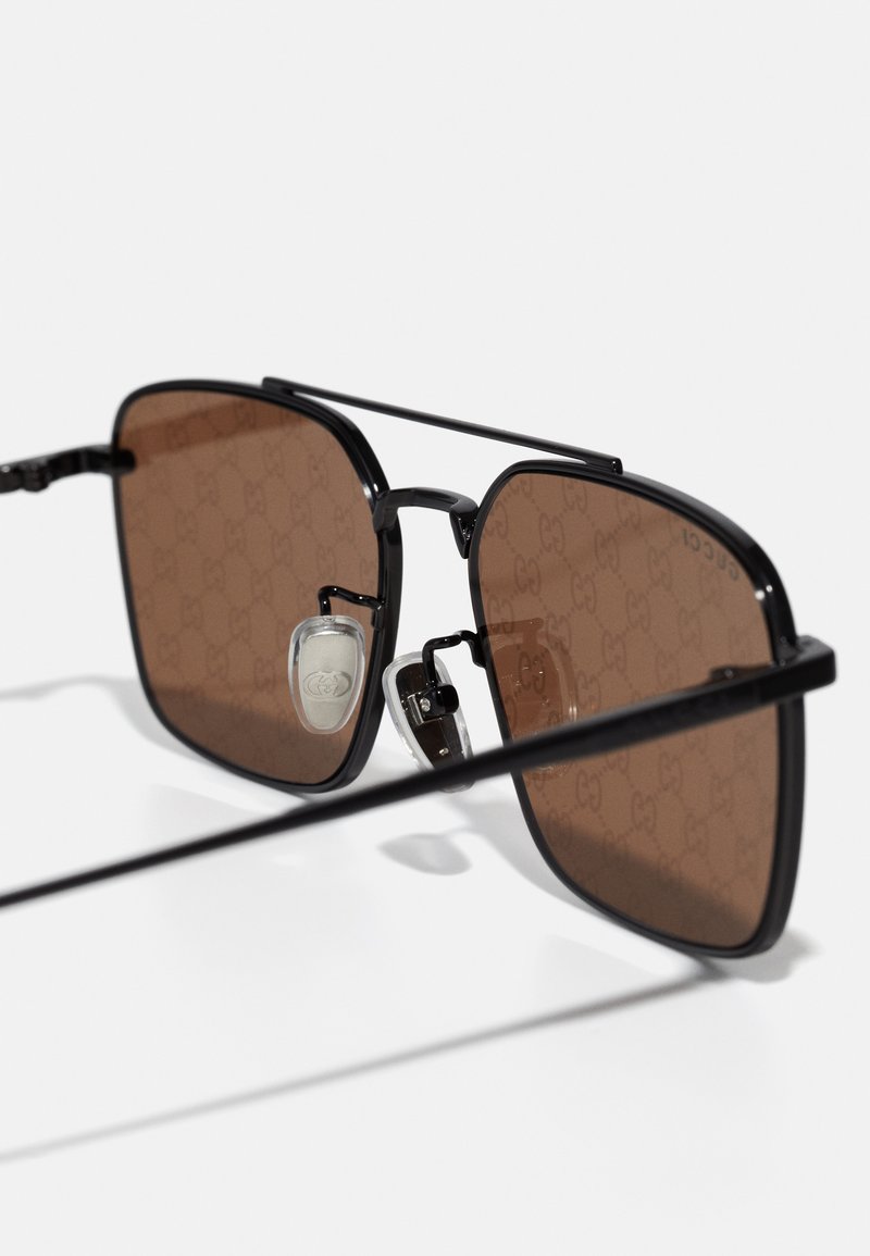 Occhiali da sole neri a forma quadrata con lenti di colore marrone, caratterizzati da un sottile motivo del logo e accenti in metallo. Inclusi supporti per il naso regolabili.