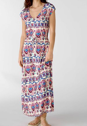 Femme portant une robe maxi sans manches à col en V avec un motif géométrique multicolore et des sandales, debout devant un fond uni.