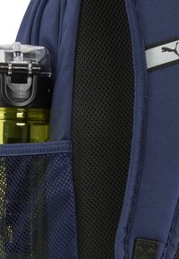 Mochila azul marino con un bolsillo lateral de malla que sostiene una botella de agua negra con tapa transparente. Tela texturizada y correas ajustables visibles.