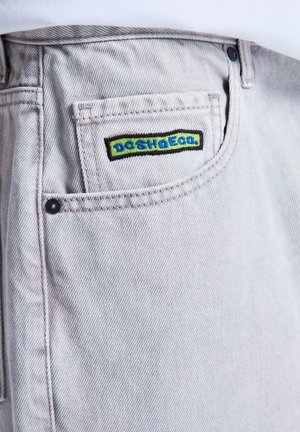 Close-up van het zakje van lichtgrijze spijkerbroek met geborduurd groen en blauw DC SHOES logo op het kleine muntzakje.