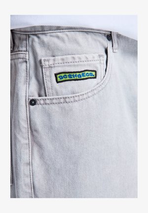 Nahaufnahme der Tasche einer hellgrauen Jeans aus Denim mit einem bestickten grünen und blauen DC SHOES Logo-Patch auf der kleinen Münztasche.