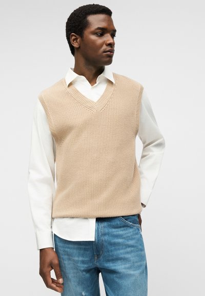 Άνδρας που φοράει μπεζ πλεκτό αμάνικο γιλέκο πάνω από λευκό πουκάμισο και μπλε τζιν, κοιτάζει στο πλάι με φόντο μονόχρωμο.