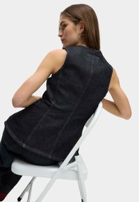 Dunkelblaues ärmelloses Oberteil mit körperbetontem Design, sichtbaren Nahtdetails und hohem Ausschnitt. Das Model sitzt auf einem weißen Klappstuhl.