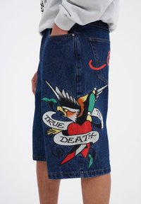 Pantalones cortos de mezclilla con un lavado azul oscuro, que presentan un colorido estampado de estilo tatuaje en la pierna izquierda con un corazón, un pájaro y una pancarta que dice "MUERTE VERDADERA".