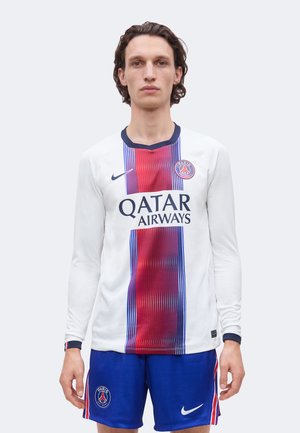 Muž v bielom futbalovom drese Paris Saint-Germain s červenými a modrými vertikálnymi pruhmi a modrými šortkami s logami tímu.