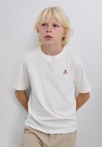 Bílé bavlněné tričko s krátkým rukávem, zdobené červenou výšivkou lebky a zkřížených kostí na hrudi. Hladká textura a klasický kulatý výstřih.