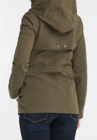 Persona che indossa una giacca con cappuccio verde oliva con dettagli a bottoni sulla patta posteriore, abbinata a jeans blu scuro, mani nelle tasche.