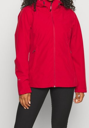 Femme portant une veste zippée rouge vif avec capuche et des leggings noirs, debout devant un fond clair uni.