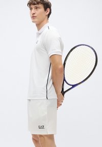 Biela polokošeľa s okrúhlym golierom s tmavomodrým pruhom. Zodpovedajúce biele šortky s logom EA7. Drží tenisovú raketu.