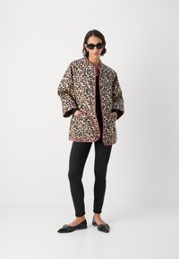 Leopardenmuster Steppjacke mit pinken Akzenten, ausgestattet mit zwei Fronttaschen, kombiniert mit schwarzen Slim-Jeans und schwarzen flachen Schuhen.