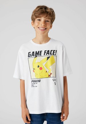 Uśmiechający się chłopiec w białej, za dużej koszulce z Pikachu i napisem „GAME FACE!”, stojący na tle jednolitego jasno szarego tła.