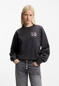 Ξανθιά γυναίκα με τα χέρια στις τσέπες φοράει μαύρο φούτερ με μικρά πολύχρωμα λουλούδια και κείμενο στο στήθος, συνδυασμένο με γκρίζα τζιν με κουμπιά.