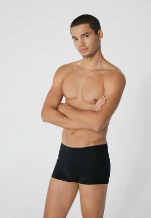 Black boxer briefs made of smooth fabric, featuring a wide waistband and a fitted design, showcased on a model with crossed arms.